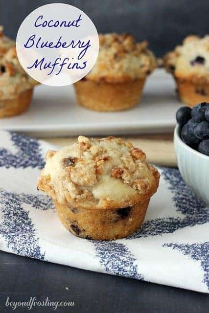 A close up of a Coconut Blueberry Muffin topped with streusel and sitting next to a bowl of fresh blueberries.