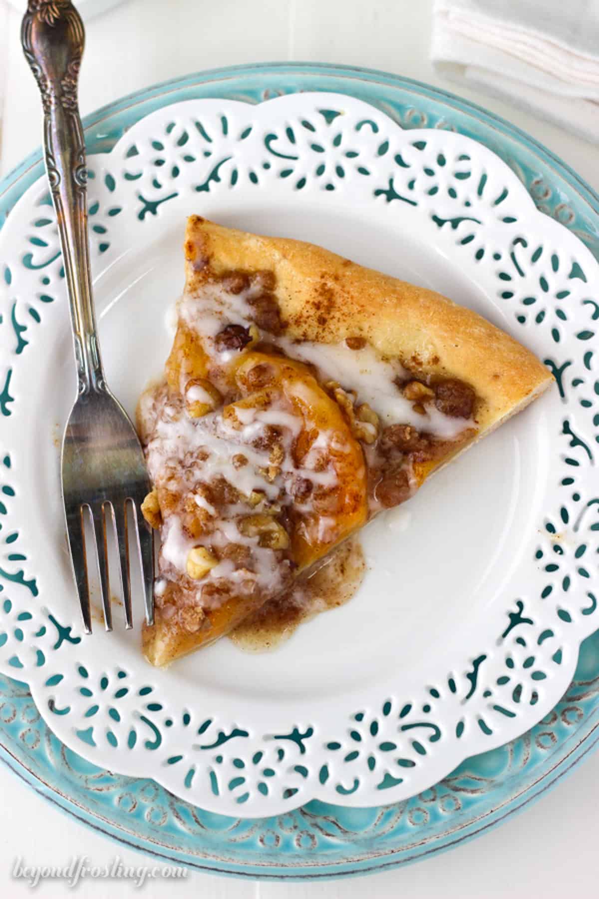 Peach pizza is a sweet dessert pizza loaded with juicy peach pie filling, buttery streusel, and walnuts, topped with a vanilla drizzle. Overhead view of a slice of peach pizza on a plate next to a fork.