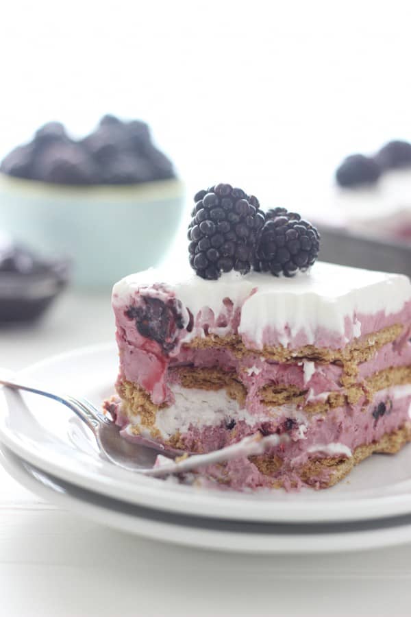 A slice of blackberry icebox cake on a double stacked white plate. It has a few bites taken out of it showing the inside of the cake, and a silver fork is resting on the plate. 