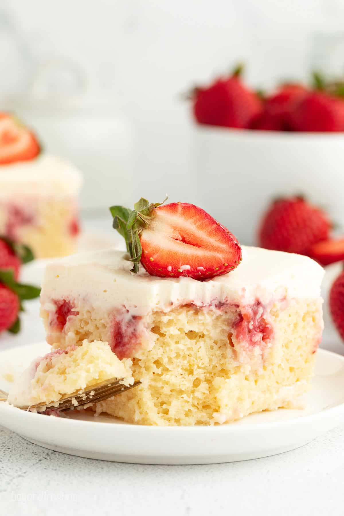 Close up of a slice of strawberry cheesecake poke cake with a forkful missing from the corner, on a white plate.
