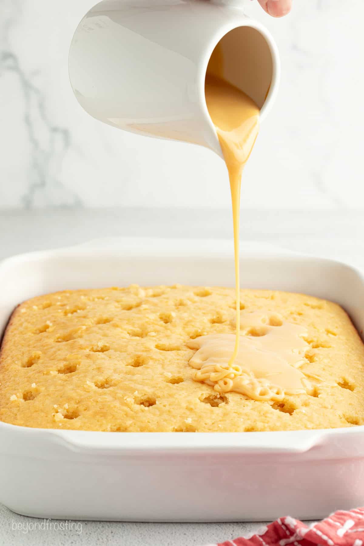 Condensed milk being poured from a ceramic jug over a vanilla cake with holes poked in the surface.