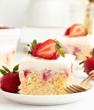 A slice of strawberry cheesecake poke cake garnished with fresh strawberries on a white plate next to a fork.