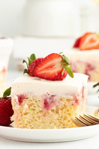 A slice of strawberry cheesecake poke cake garnished with fresh strawberries on a white plate next to a fork.
