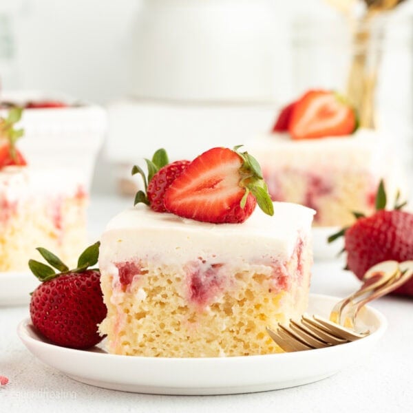 A slice of strawberry cheesecake poke cake garnished with fresh strawberries on a white plate next to a fork.