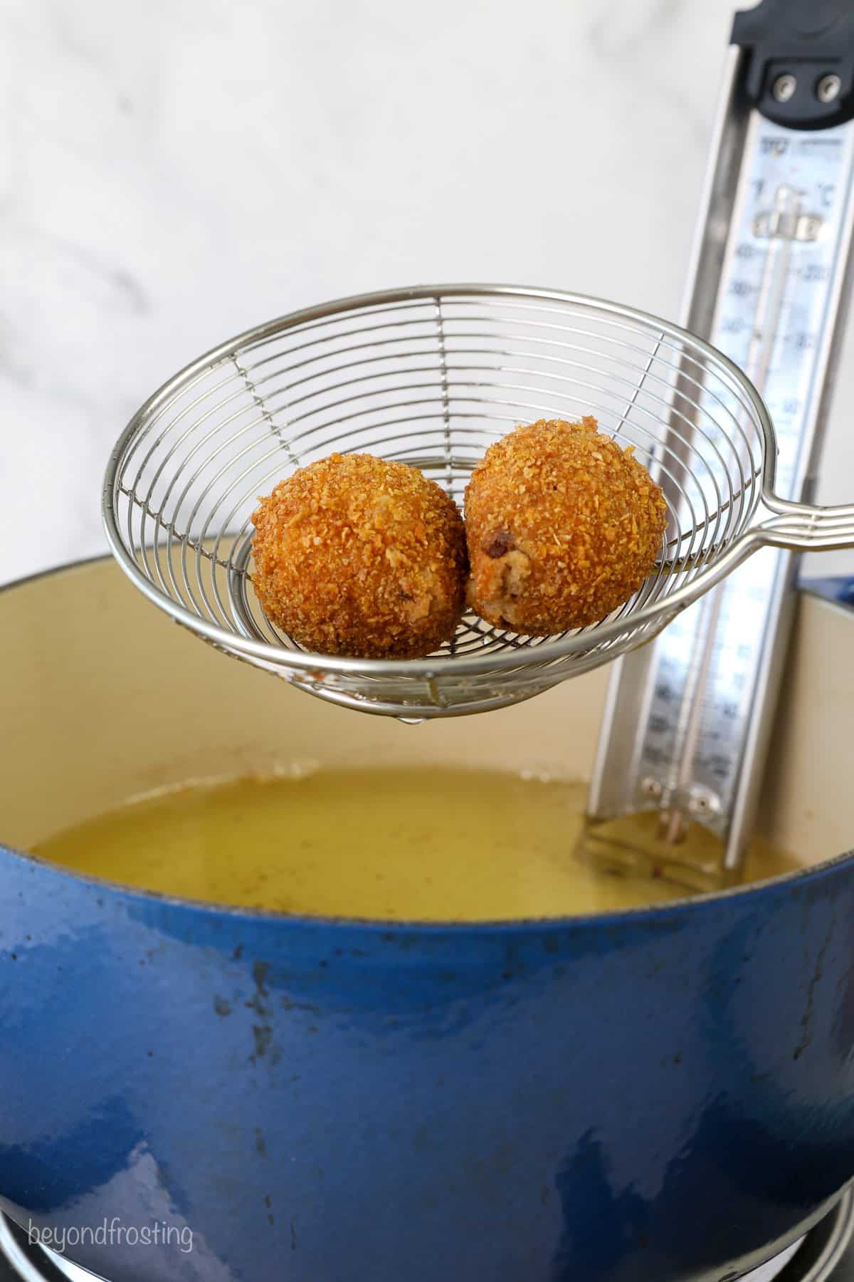 Two deep fried cookie dough balls in a metal slotted spoon, held over a pot of hot oil fitted with a candy thermometer.