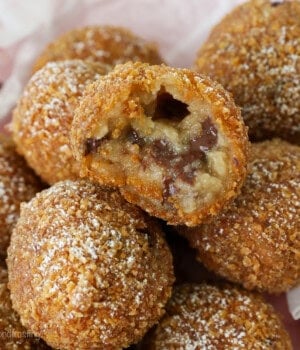 Deep fried cookie dough balls stacked in a paper-lined basket, with a bite missing from the top ball.