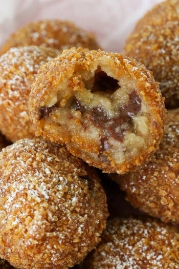 Deep fried cookie dough balls stacked in a paper-lined basket, with a bite missing from the top ball.