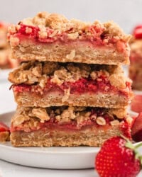 A stack of three strawberry rhubarb bars on a white plate next to a fresh strawberry.