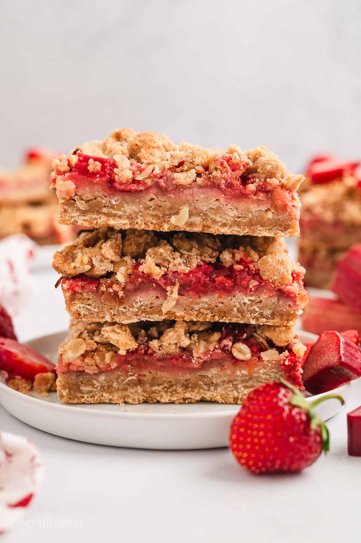 A stack of three strawberry rhubarb bars on a white plate next to a fresh strawberry.