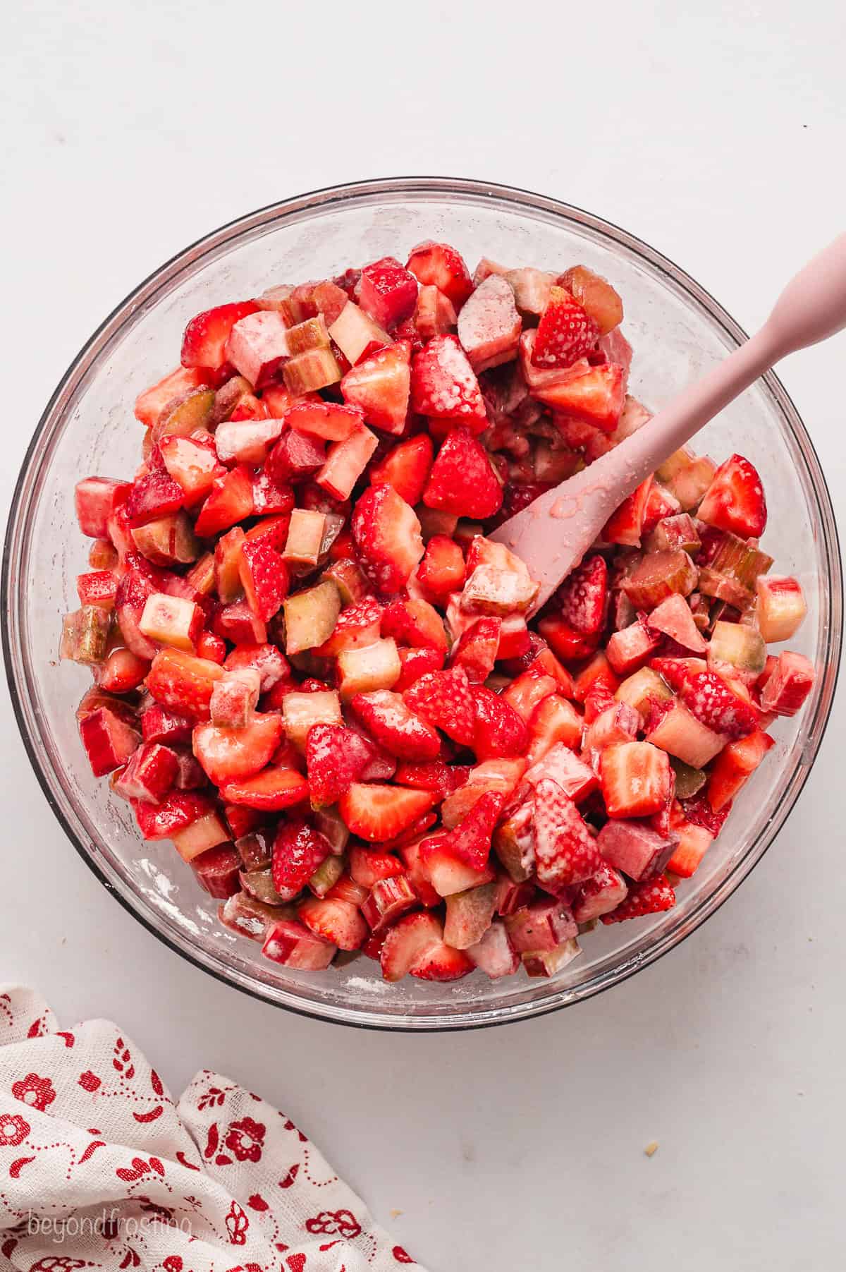 Diced strawberries and rhubarb combined in a glass bowl with a wooden spoon.