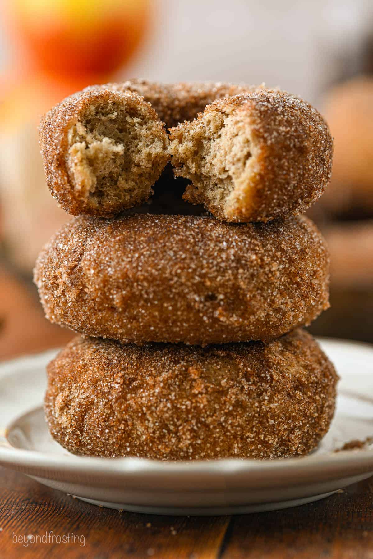 Three apple cider donuts stacked on top of one another on a white plate, with a bite missing from the top donut.