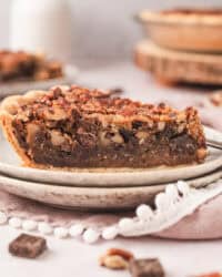 A slice of bourbon chocolate pecan pie on a plate with the rest of the pie in the background.