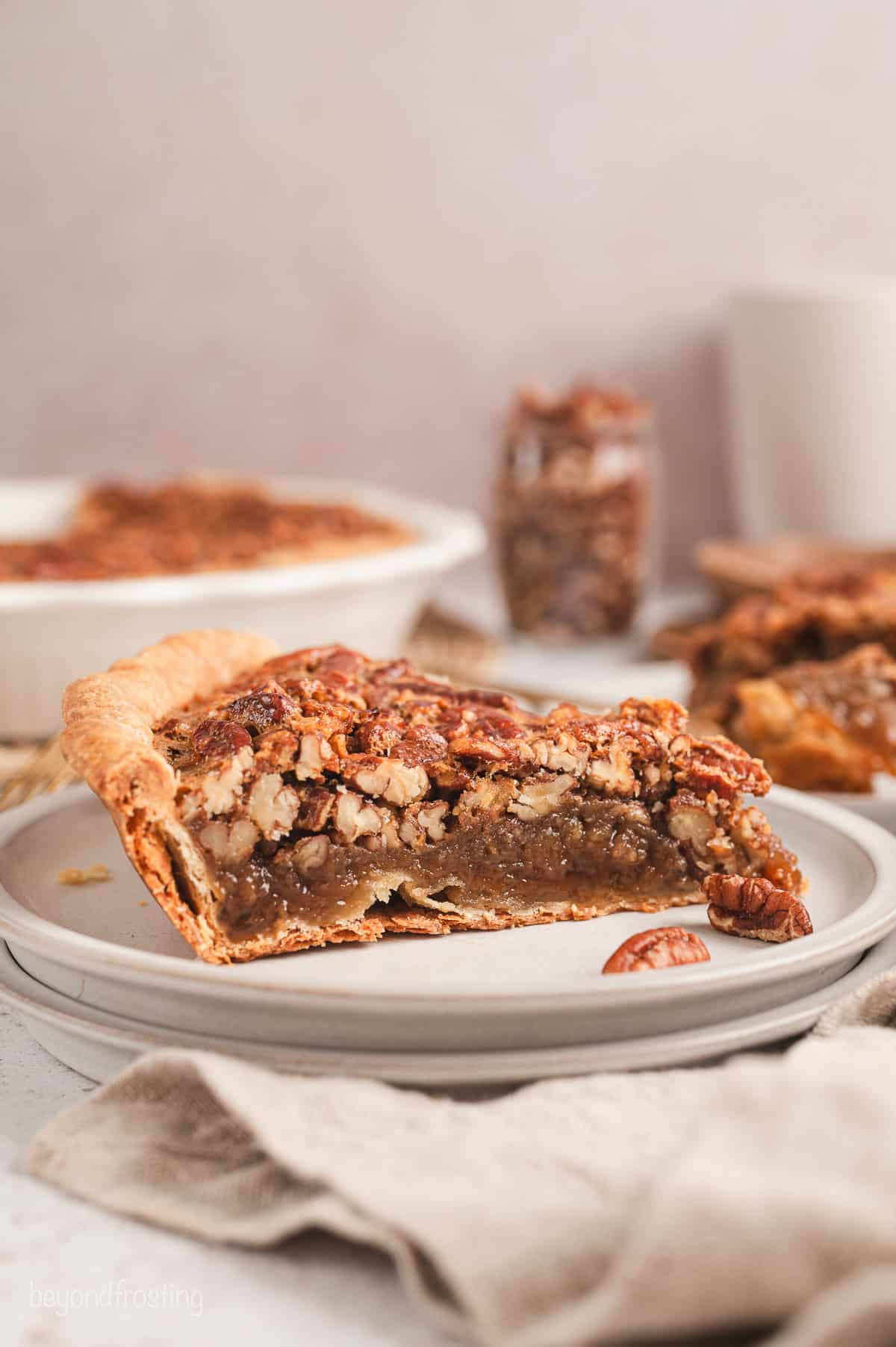 A slice of pecan pie on a plate with the rest of the pie in the background.