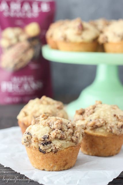 Three maple pecan banana muffins on a piece of parchment paper with more muffins on a cake stand in the background