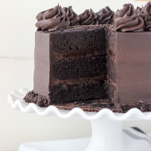 A chocolate layer cake sitting on a white scalloped edge cake plate with a big slice missing. The cake is decorated with chocolate rosettes and sprinkles