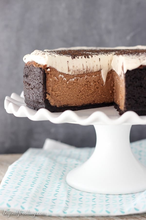 A big chocolate cheesecake on a white ruffled edge cake stand with a couple of slices missing