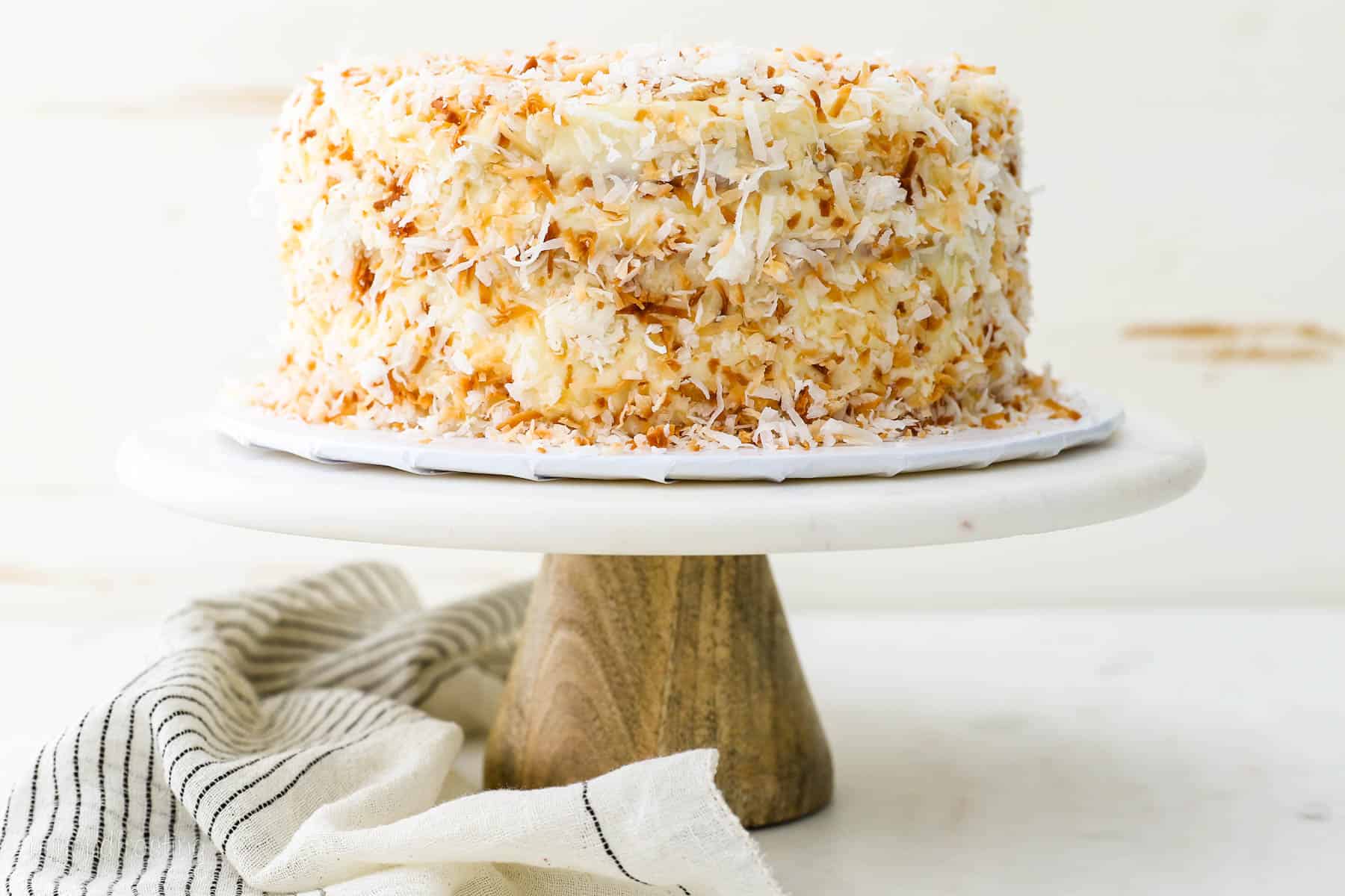 Coconut custard cake covered with toasted coconut on a white cake stand with a wooden base.