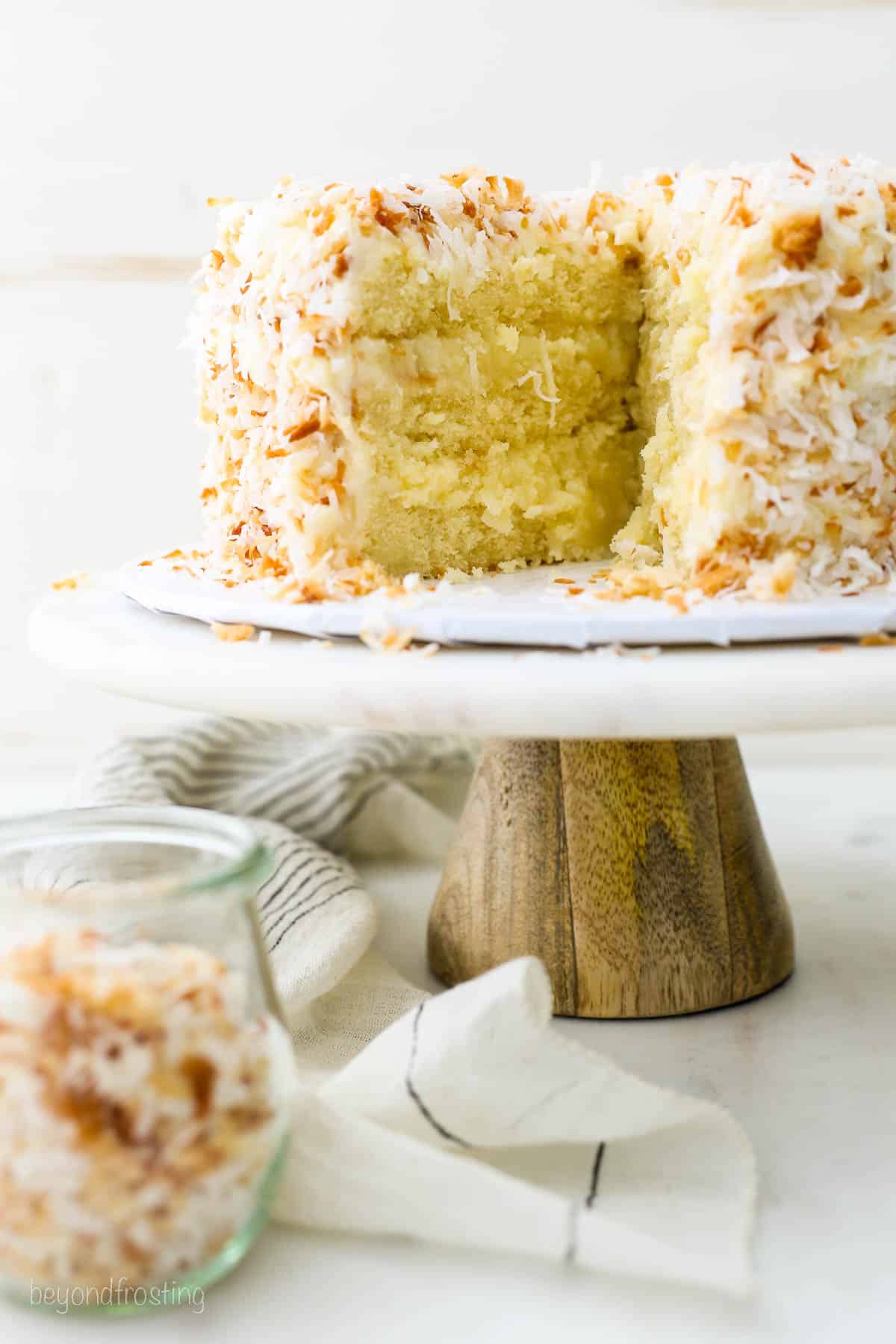 Coconut custard cake covered with toasted coconut with a slice missing, on a white cake stand with a wooden base, next to a jar of toasted coconut.
