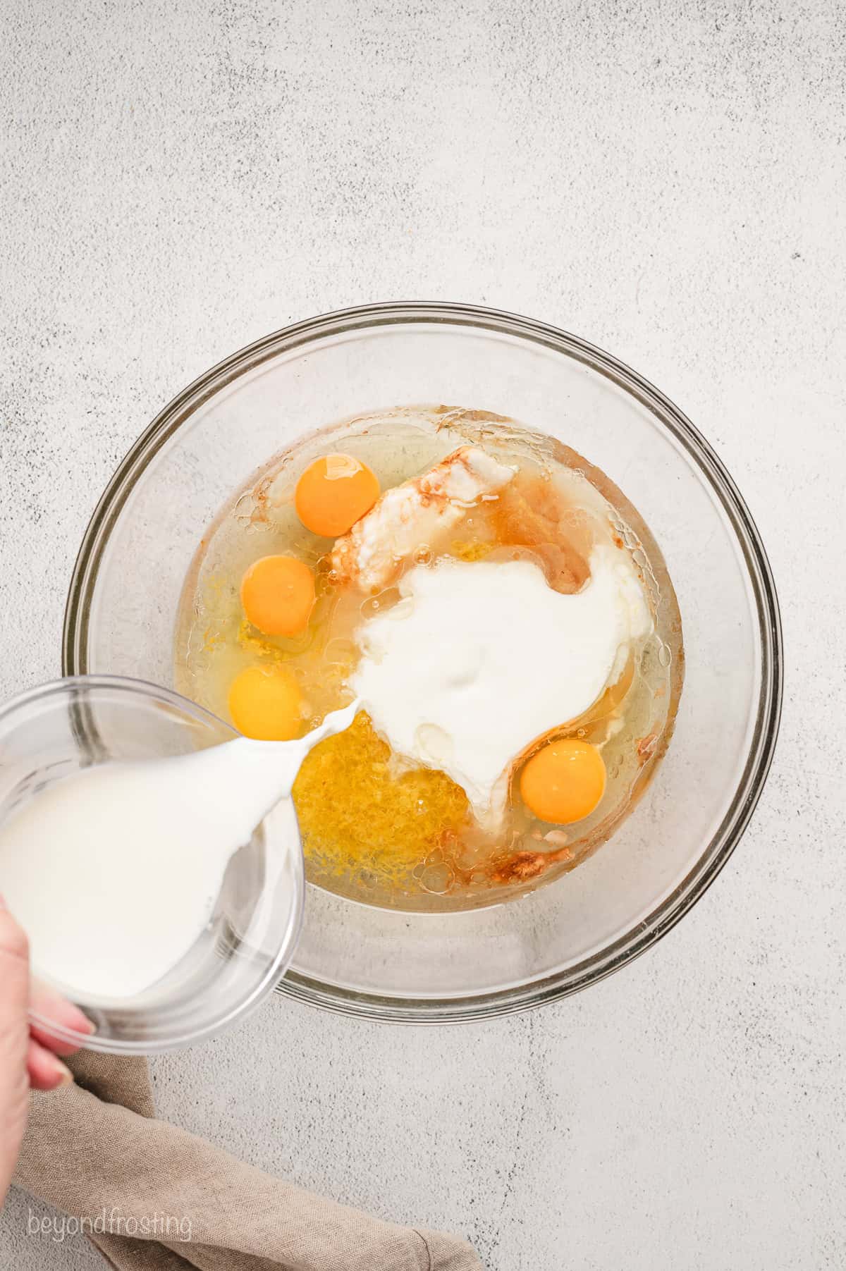 Milk being poured over eggs and wet ingredients in a glass bowl.