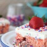 A slice of Funfetti Strawberry Poke Cake: a strawberry cake with sprinkles and topped with a whipping cream topping, sprinkles, and a large strawberry.