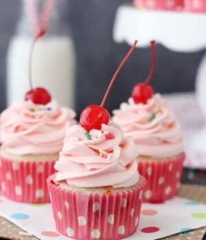 Three Cherry Almond Funfetti Cupcakes in pink cupcake wrappers and topped with a pink cherry almond frosting and a maraschino cherry.