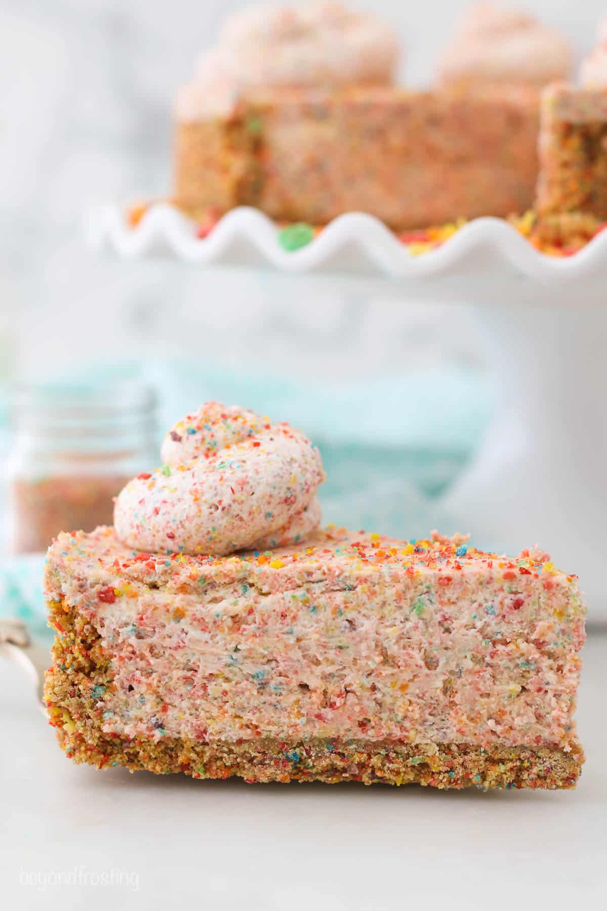 A slice of fruity pebbles cheesecake in front of a cake stand