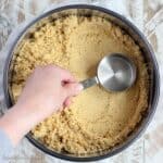 A hand using a measuring cup to press the Oreo crumbs into a springform pan