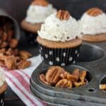 A maple pecan cupcake balancing between the divots of a cupcake tin with a bunch of whole pecans beside it