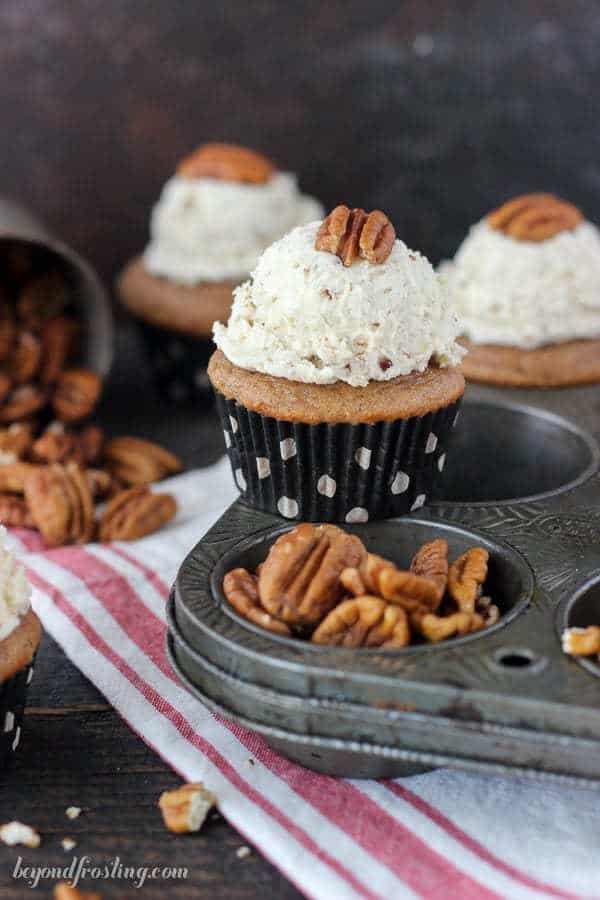 A maple pecan cupcake balancing between the divots of a cupcake tin with a bunch of whole pecans beside it