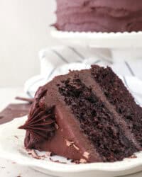 A slice of frosted chocolate sweet potato cake on a white plate with the rest of the cake on a cake stand in the background.