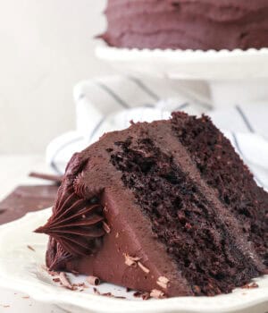 A slice of frosted chocolate sweet potato cake on a white plate with the rest of the cake on a cake stand in the background.