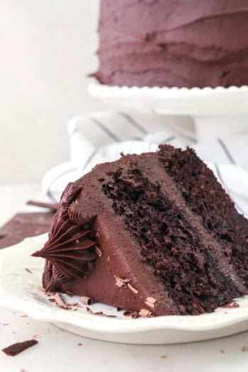 A slice of frosted chocolate sweet potato cake on a white plate with the rest of the cake on a cake stand in the background.