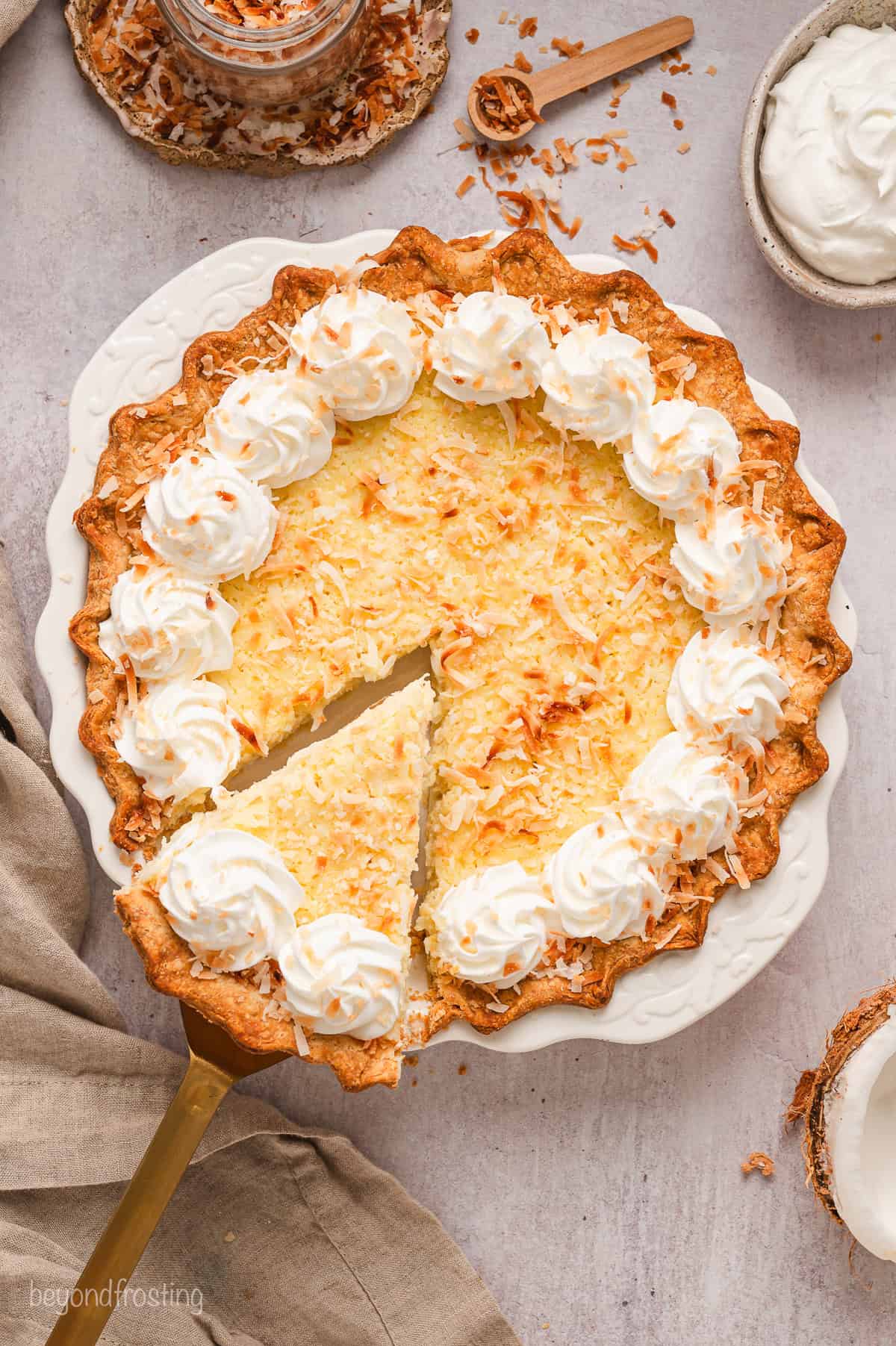 Overhead view of coconut custard pie in a pie plate decorated with a border of piped whipped cream swirls, with a slice being lifted from the pie.