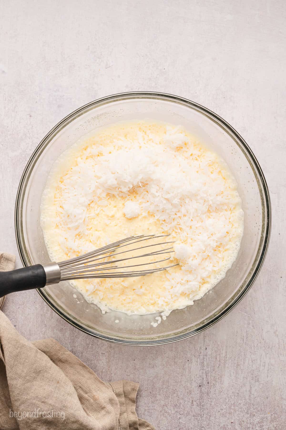 Shredded coconut added to a bowl of coconut custard, with a whisk.