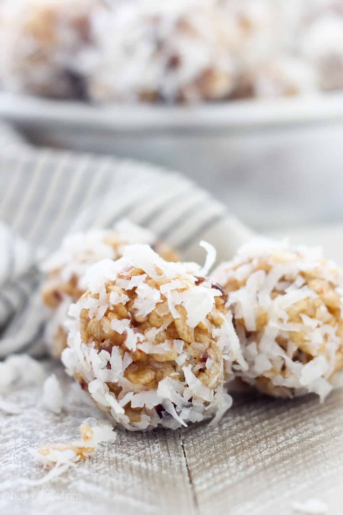 Close up of two coconut date balls on a tabletop with a plate of date balls in the background.