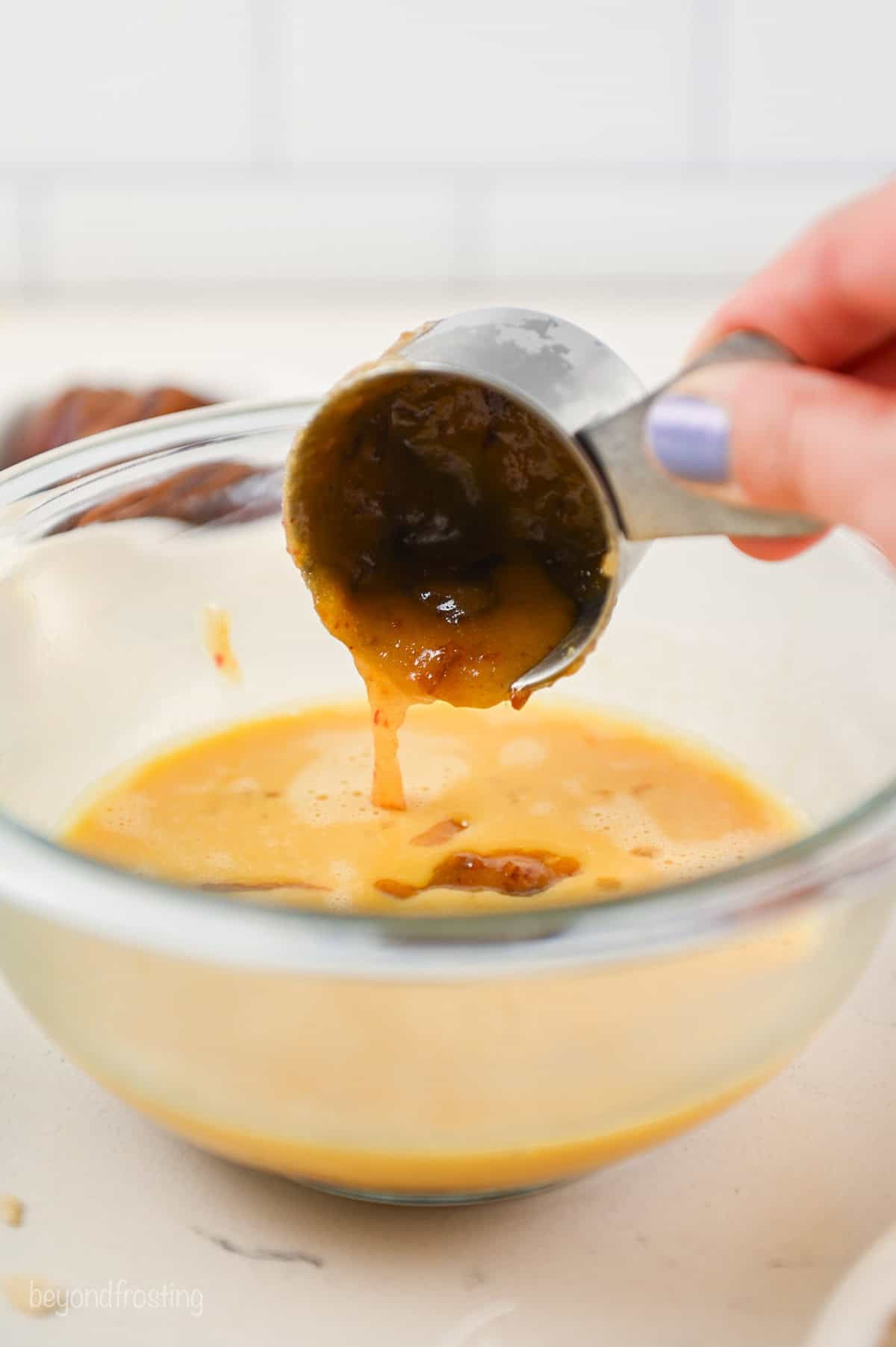 A hand tipping a small scoop of hot date mixture into a bowl of beaten egg mixture.