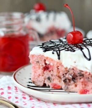 A slice of Cherry Chocolate Chip Poke Cake: a sweet cherry cake with chocolate chips, soaked in sweetened condensed milk and topped with whipped cream, a maraschino cherry, and chocolate sauce.