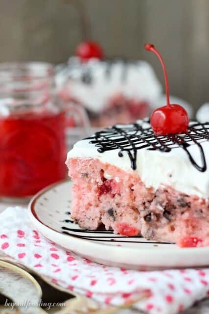 A slice of Cherry Chocolate Chip Poke Cake: a sweet cherry cake with chocolate chips, soaked in sweetened condensed milk and topped with whipped cream, a maraschino cherry, and chocolate sauce.