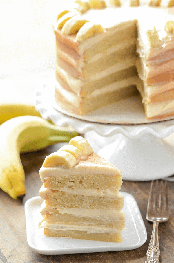 A Slice of Banana Dream Cake on a Plate Next to The Whole Layer Cake on a Cake Stand