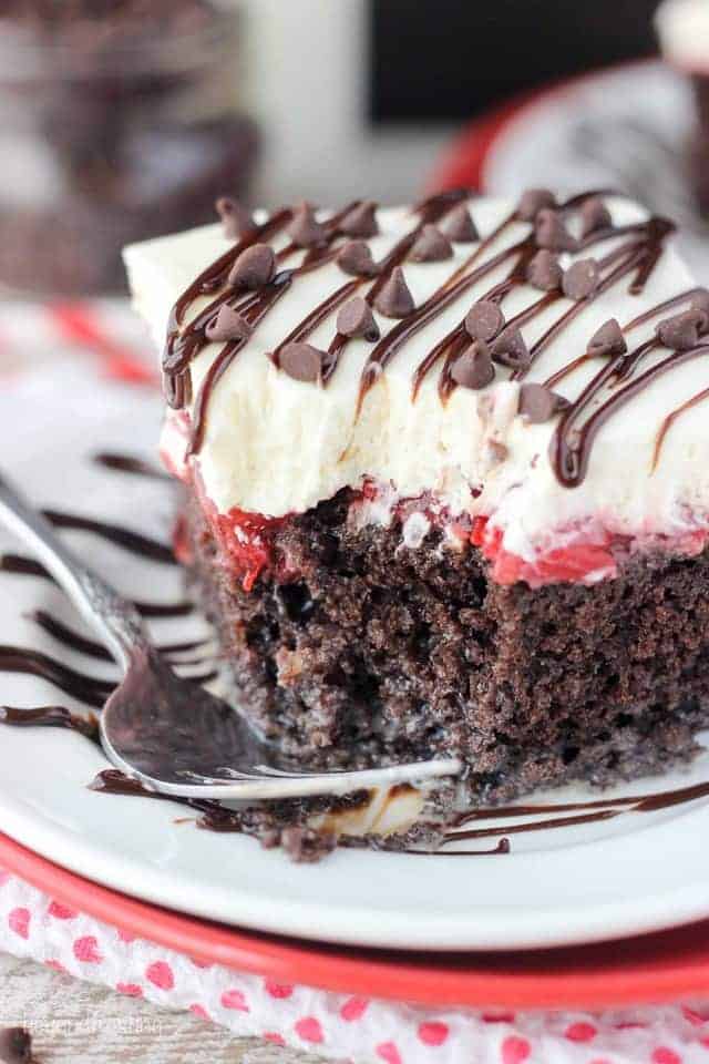A close up of a piece of Chocolate Cherry Poke Cake on a white plate with a fork and a bite taken out
