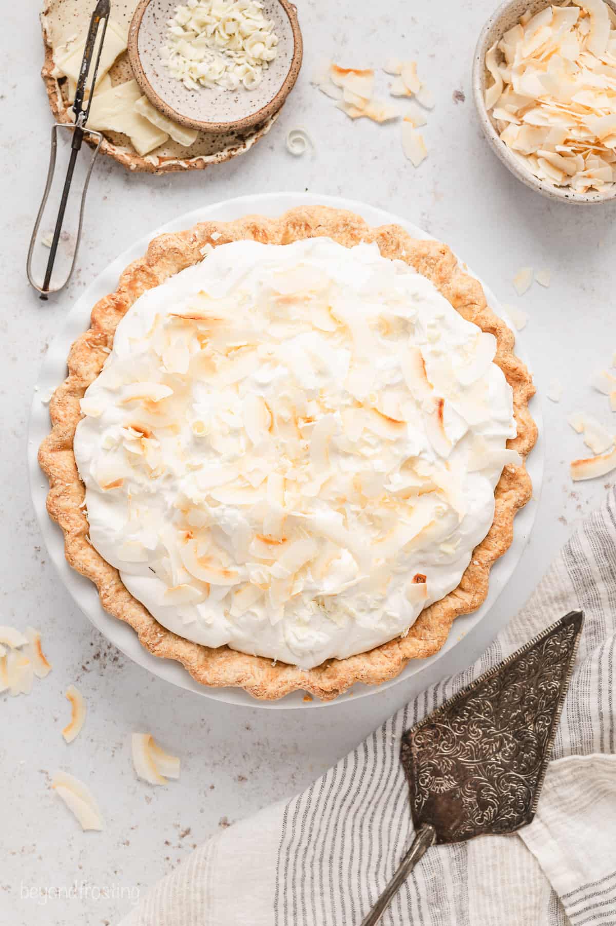 Overhead view of a whole coconut cream pie in a pie plate, next to a cake server and a bowl of coconut flakes on a countertop.