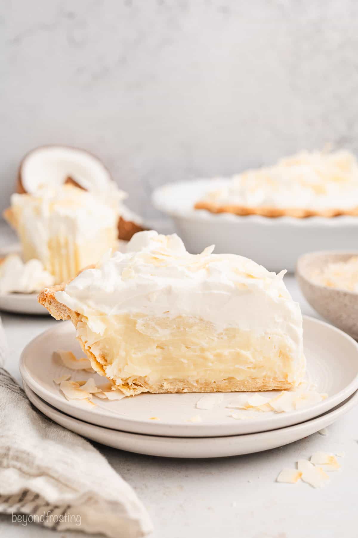 Side view of a slice of coconut cream pie on a plate, with the rest of the pie in the background.