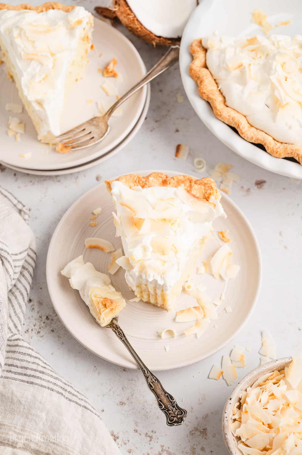 Overhead view of a slice of coconut cream pie on a plate, with a forkful missing from the end, next to a second slice on a plate and the rest of the pie in a pie plate.
