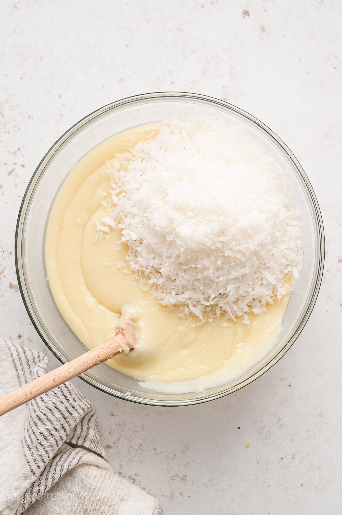 Coconut flakes added to coconut cream pie filling in a glass bowl with a wooden spoon.