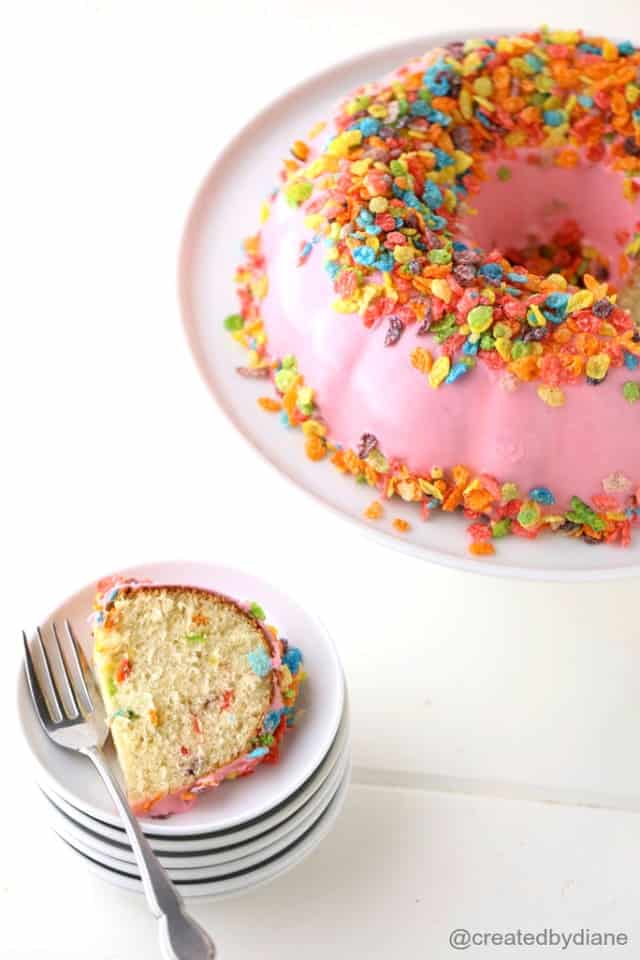 A Slice of Fruity Pebbles Cake on a Plate Next to the Full Cake on a Cake Stand