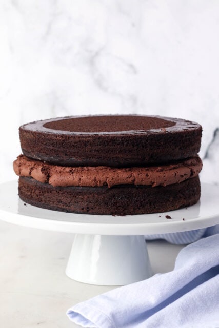 Two chocolate zucchini cake layers stacked on a cake stand, with chocolate frosting between the layers.