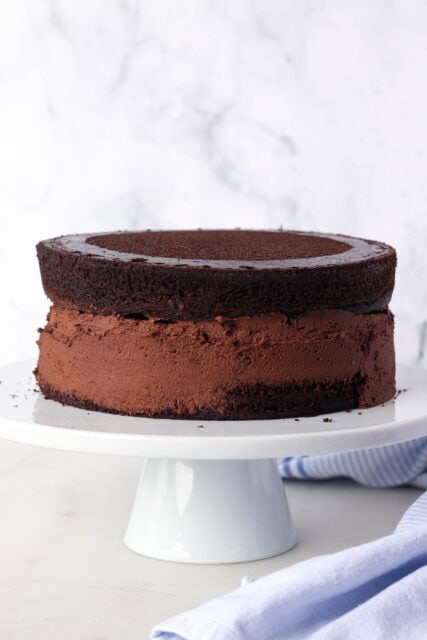 Two chocolate zucchini cake layers stacked on a cake stand, with chocolate frosting spread over the edge of cake.