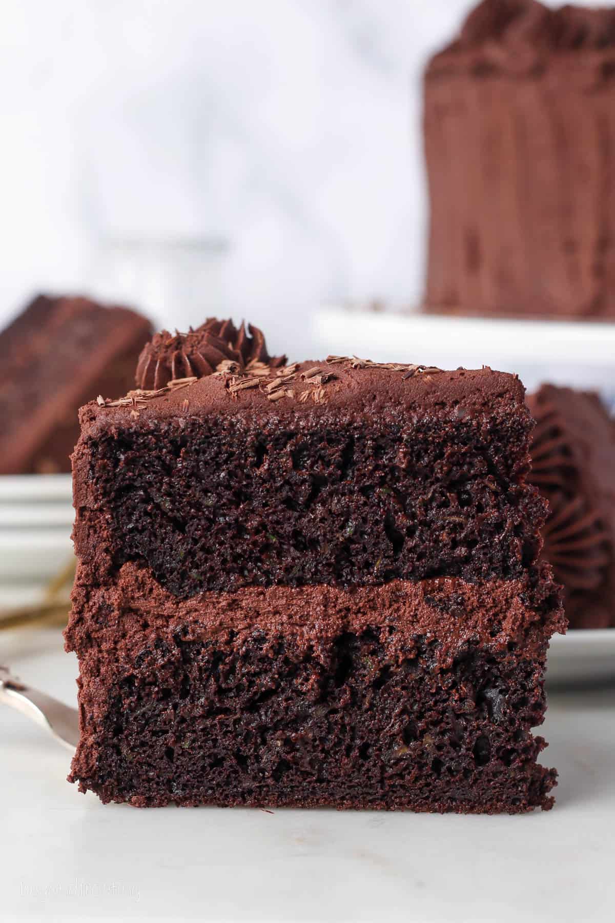 A slice of frosted chocolate zucchini cake standing upright on a plate.