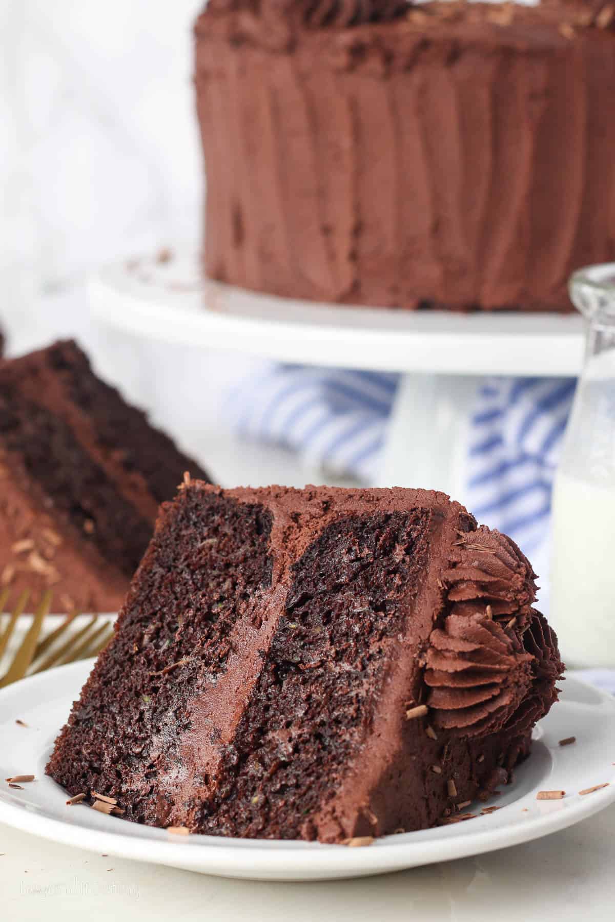 A slice of frosted chocolate zucchini cake laying on a plate, with the rest of the cake on a cake stand in the background.