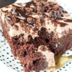 A crumbling slice of Brownie Batter Poke Cake. A rich chocolate Cake filled with chocolate pudding and topped with a brownie batter mousse and brownie batter glaze.